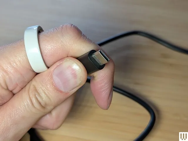 Una mano sostiene un cable con conector USB-C negro sobre una mesa de madera.